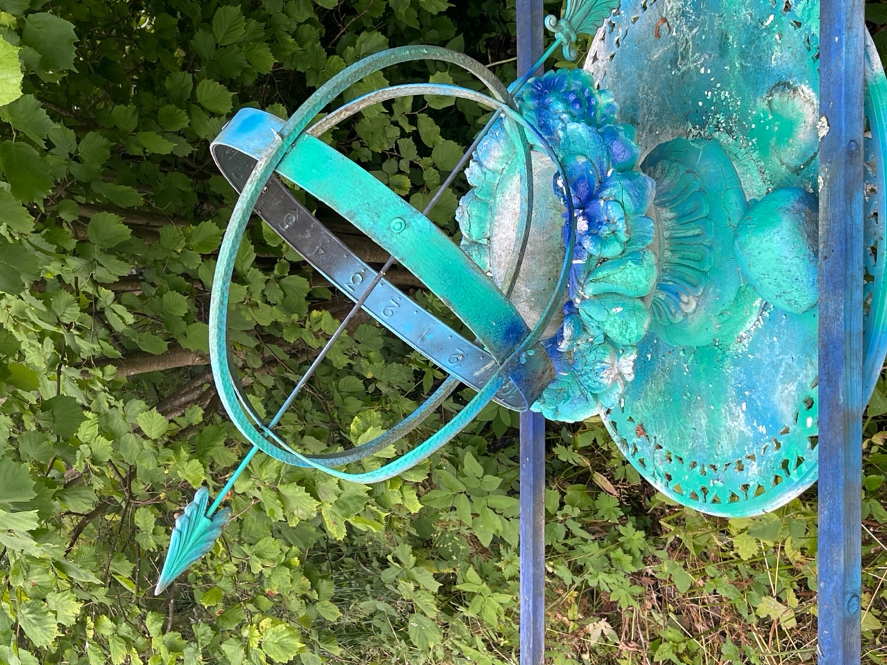 Grön-blå installation av ett gammalt solur och en blom-urna på ett gjutet stålbord